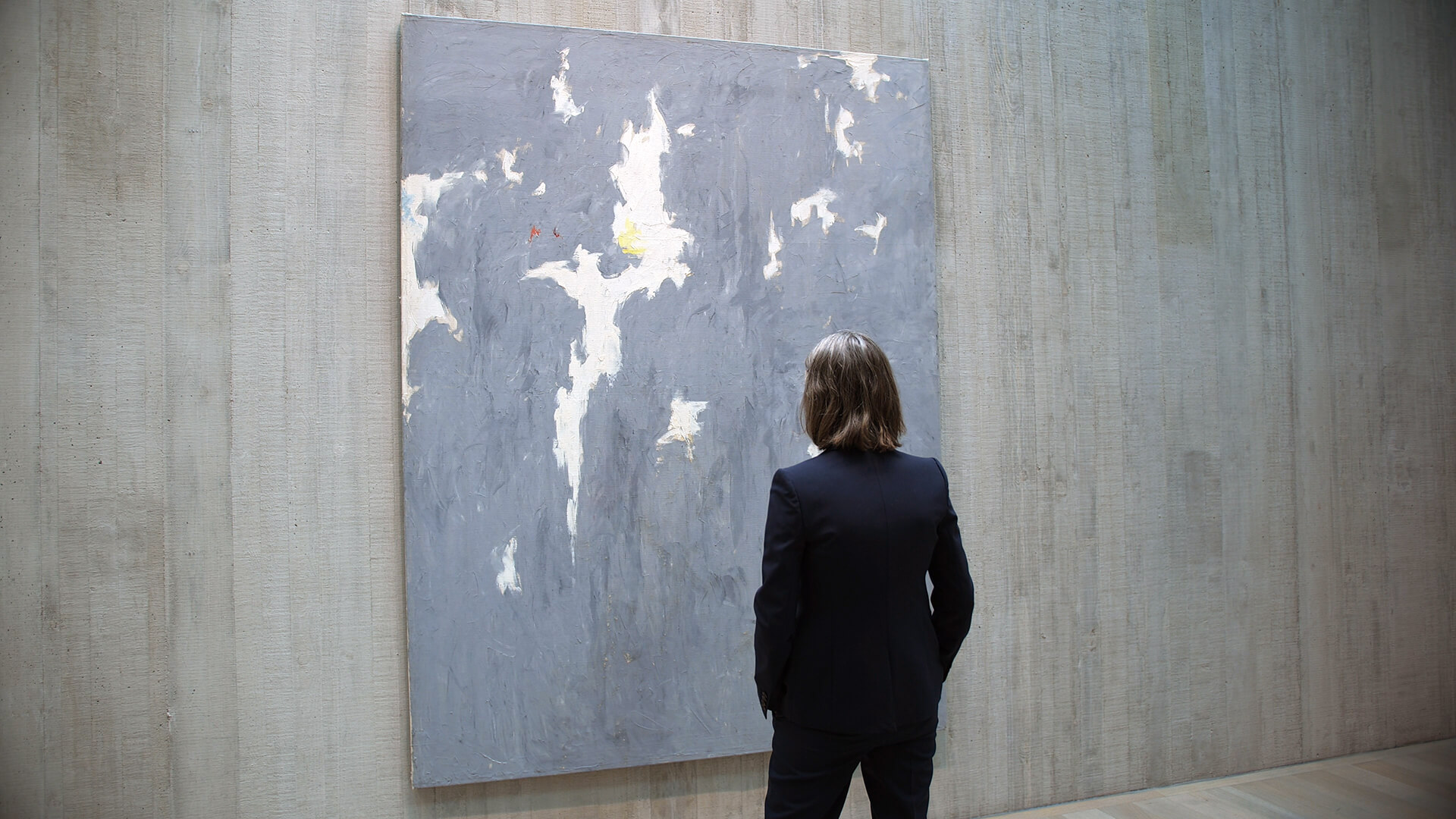 A woman looks at a blue gray painting on a concrete wall.