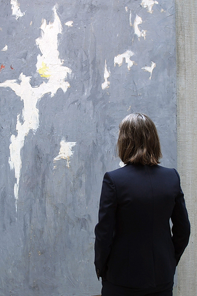 A woman wearing a dark navy blue suit stands in front of a grayish blue painting on a concrete wall