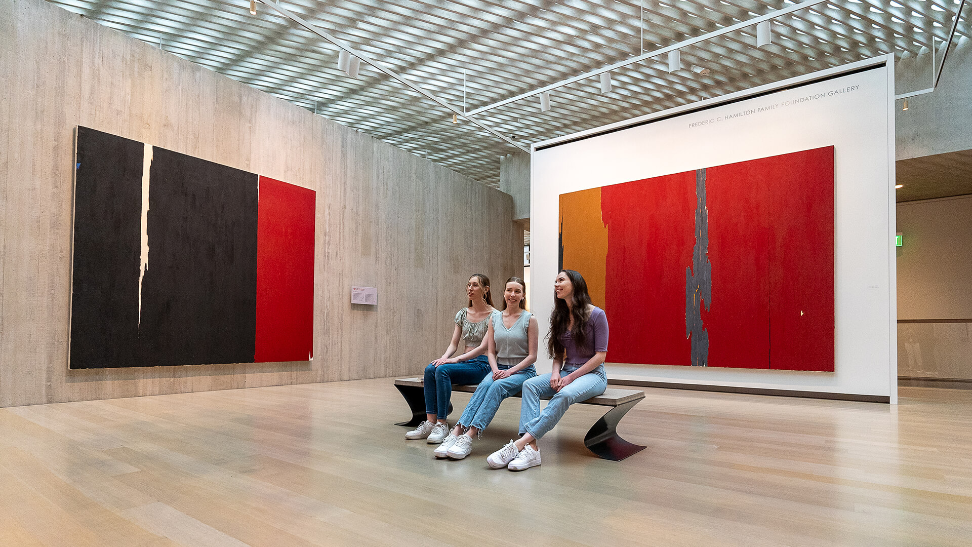 Three young women sit on a bench and look at art