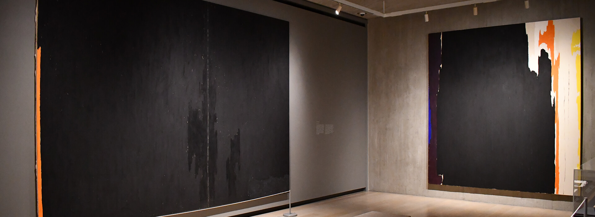 Installation image of a gallery with two abstract paintings with mostly black paint and hints of color