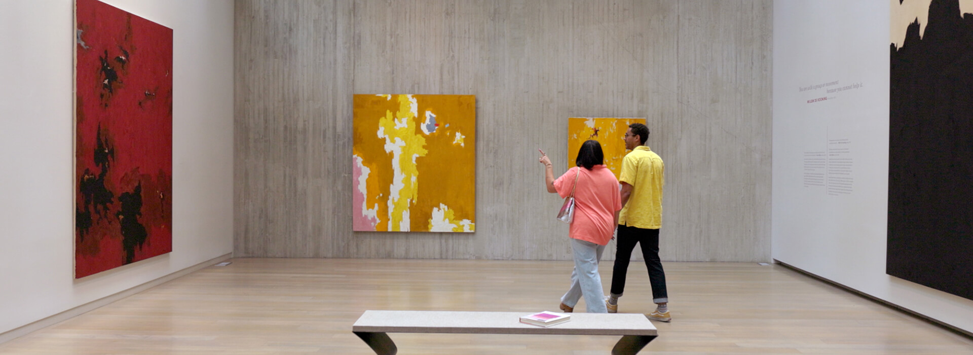 A couple wearing yellow and pink shirts walks towards a yellow and pink painting