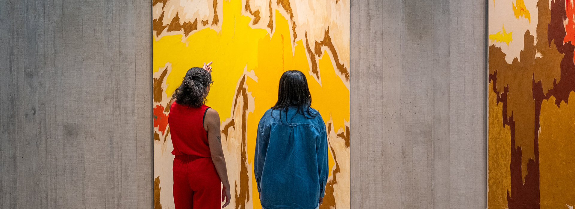 Two women look at a large yellow and orange painting while one of them points at it.