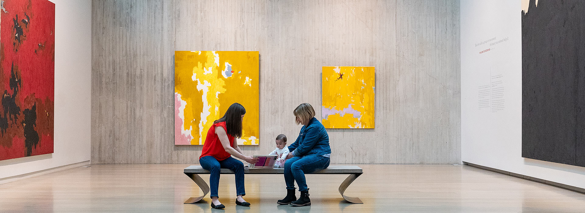 A grandmother and mother sit with a baby on a bench in an art gallery and show her a book