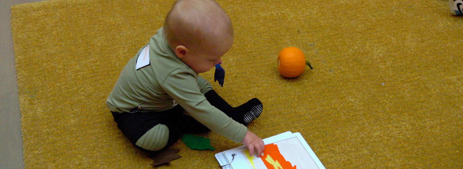 A baby sits on the floor and touches an orange and yellow shape in a tactile book