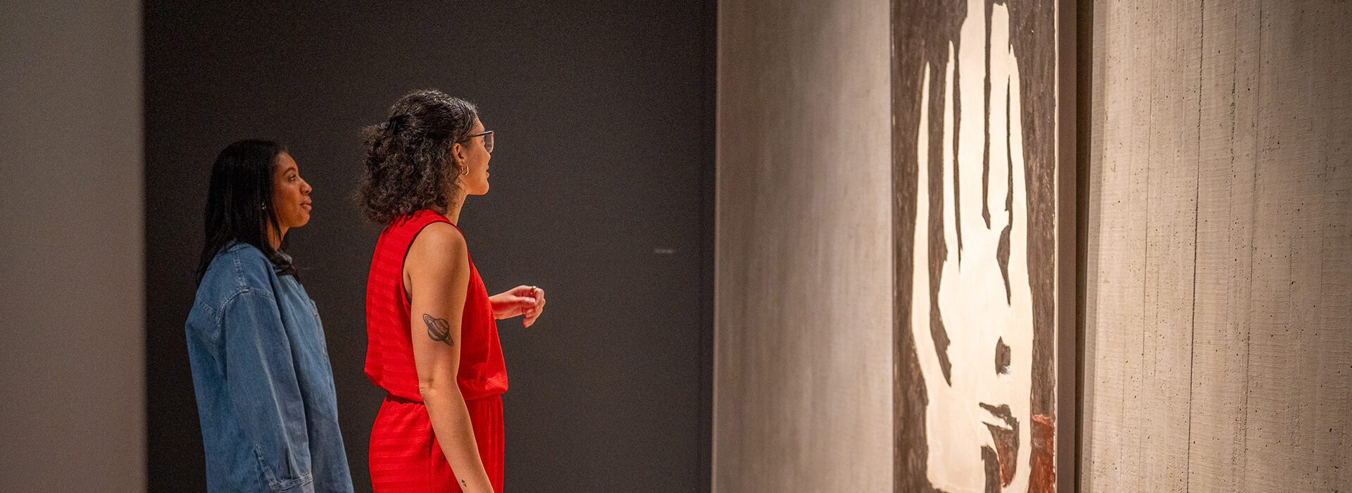 Two women look at an abstract black and white painting in a darkly lit gallery