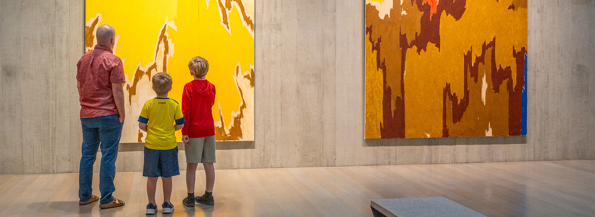 A father and his two sons look at a large abstract yellow and gold painting