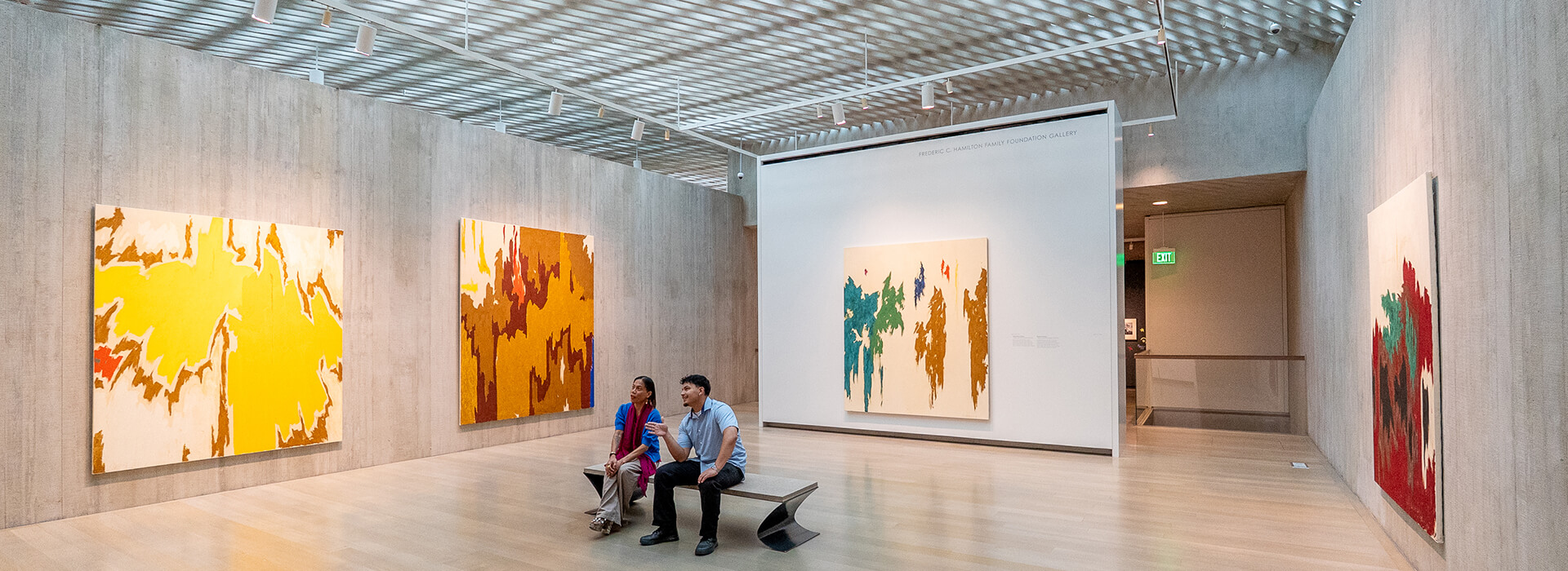 Two people sit on a bench in a large gallery with colorful abstract paintings