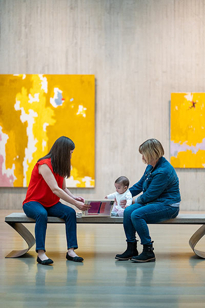 A grandmother and mother sit with a baby on a bench in an art gallery and show her a book