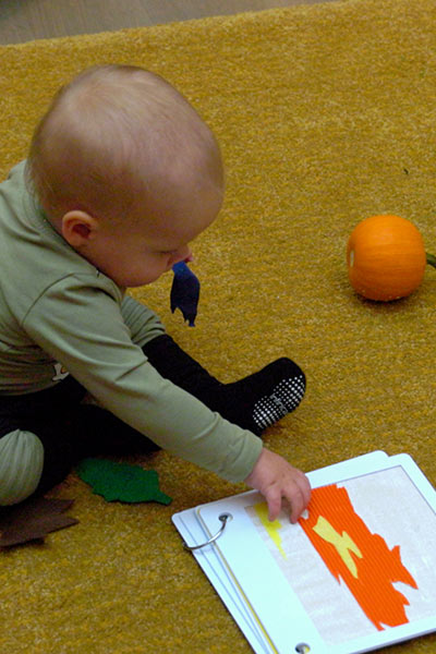 A baby sits on the floor and touches an orange and yellow shape in a tactile book