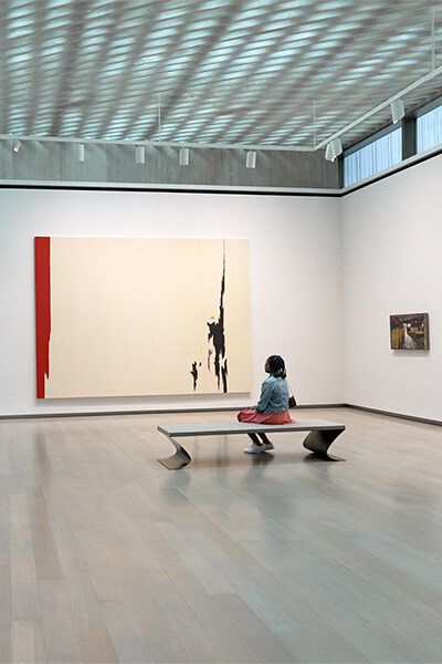 A young woman sits on a bench and looks at large abstract paintings on the wall