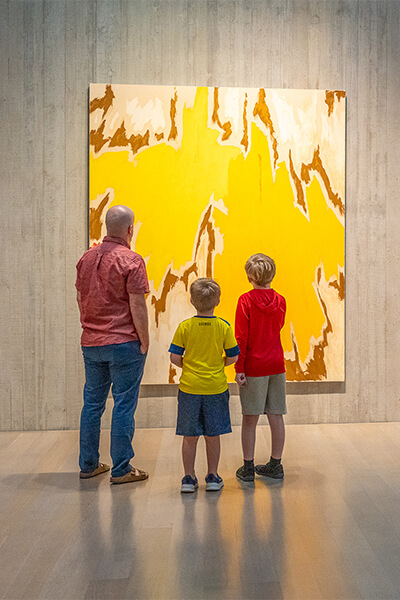 A father and his two sons look at a large abstract yellow and gold painting