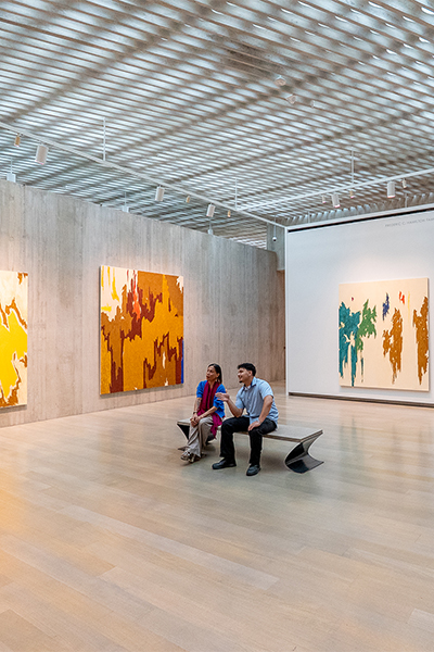 Two people sit on a bench in a large gallery with colorful abstract paintings