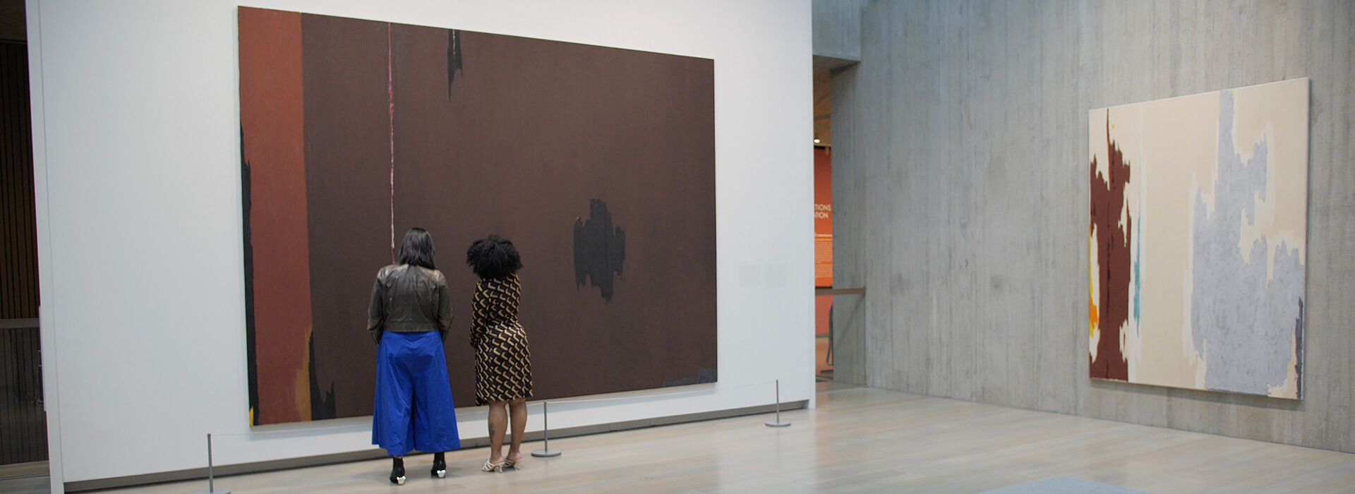 Two women look at a large abstract painting with brown, black, and ochre paint