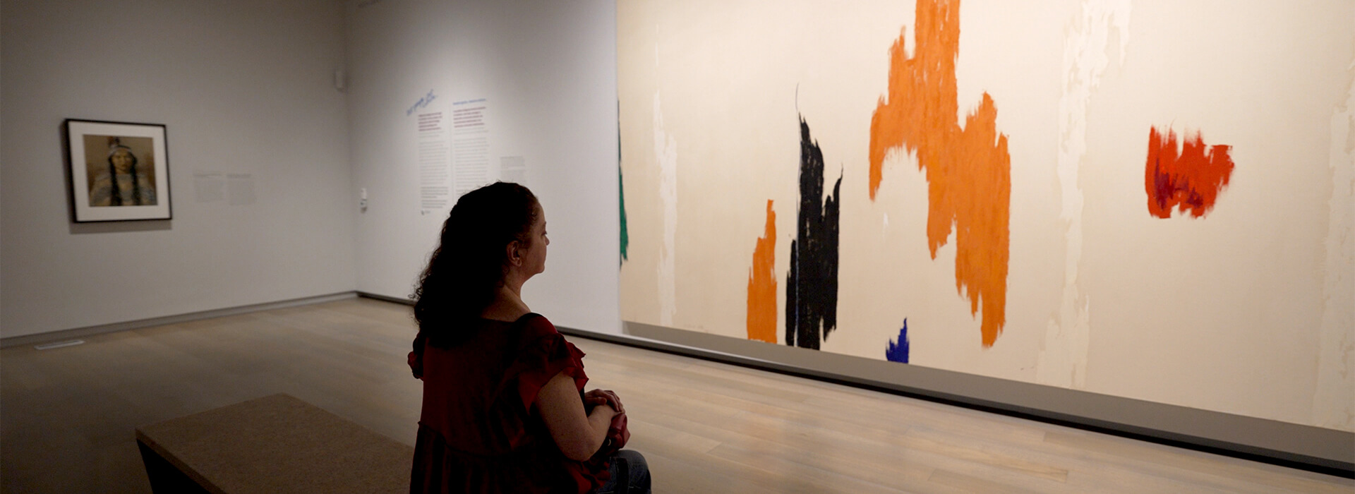 A museum visitor sits on a bench and looks at a large abstract painting.