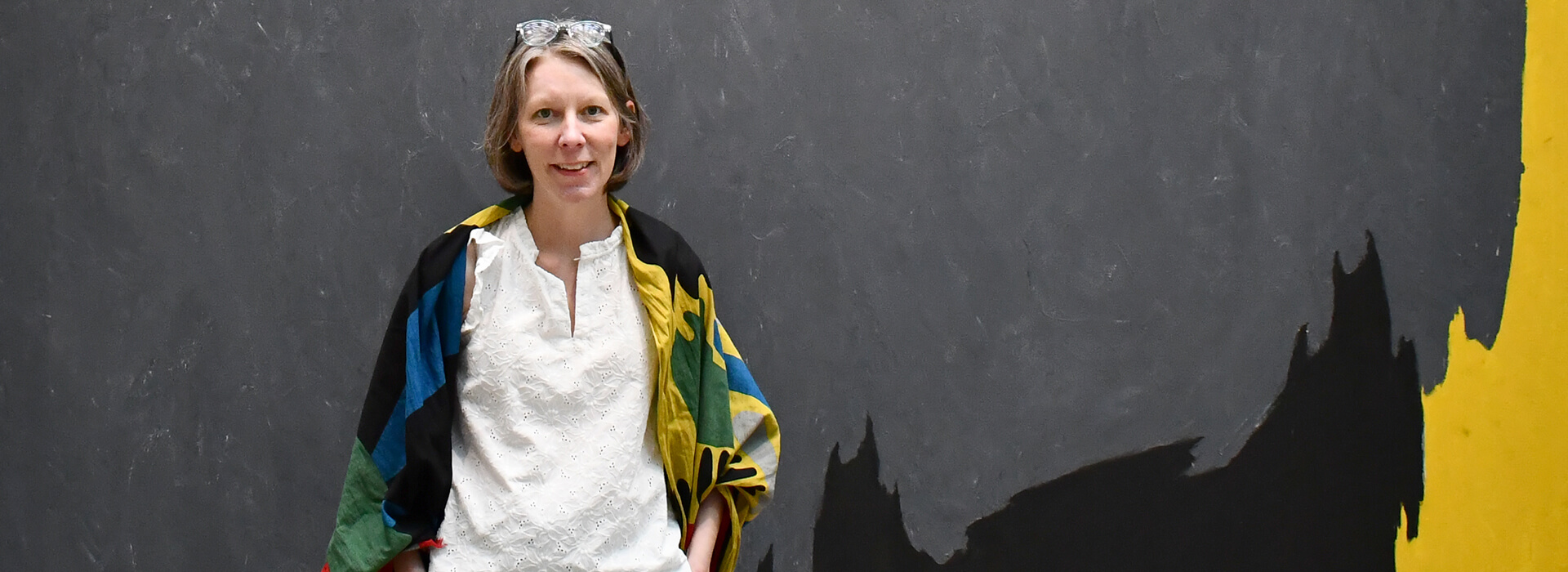 A woman stands in front of a large gray painting with some yellow, black and red around the edge