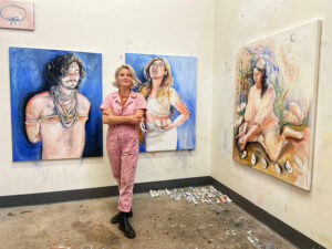 An artists wears a pink jumpsuit and stands with her arms crossed in front of large paintings of people