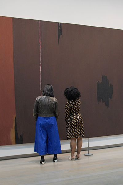 Two women look at a large abstract painting with brown, black, and ochre paint