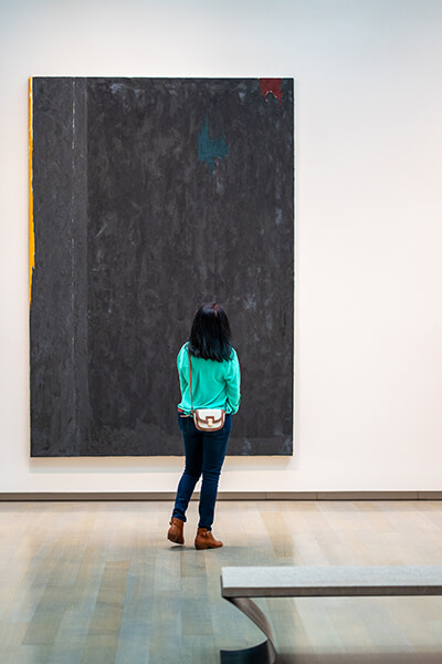 A woman stands in front of a large black painting with hints of gray, orange, red, and green