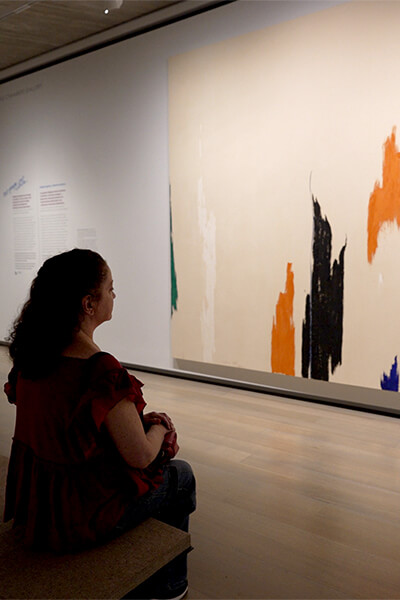A museum visitor sits on a bench and looks at a large abstract painting.