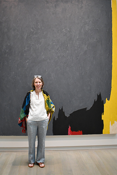 A woman stands in front of a large gray painting with some yellow, black and red around the edge