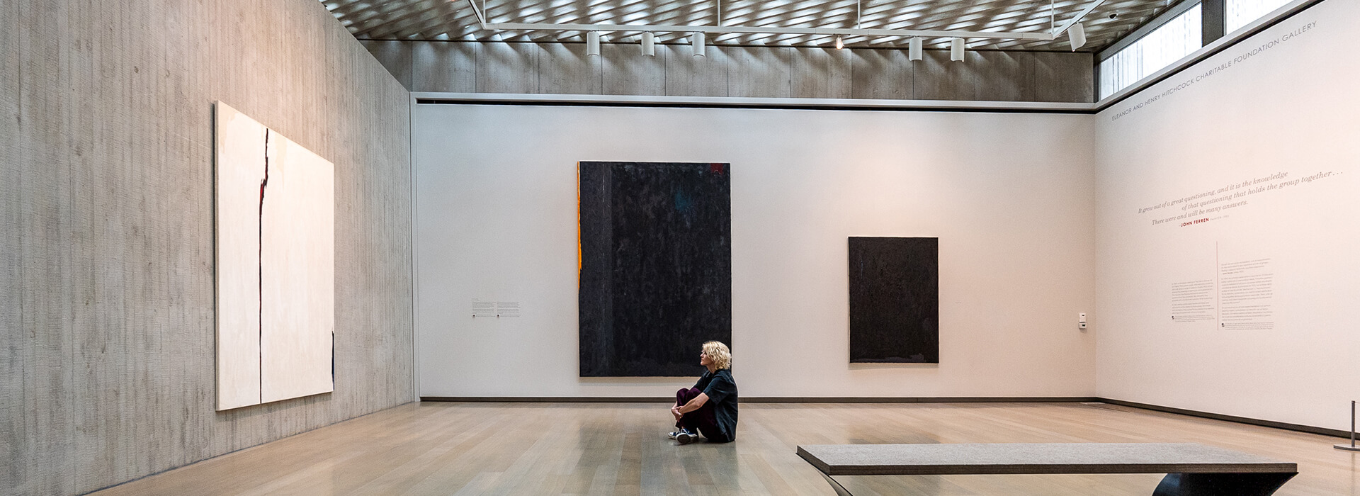 A young woman sits on the floor of a gallery and looks up at a large white abstract painting