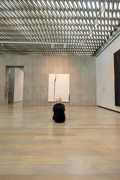 A young woman sits on the floor of a gallery and looks up at a large white abstract painting