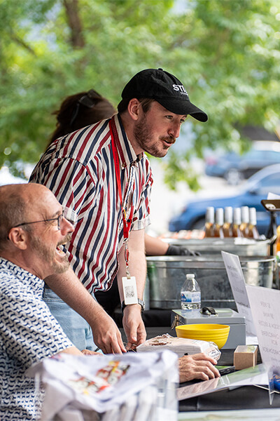 Two men talk to people while sitting and standing behind a booth