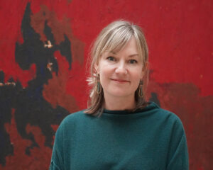 A woman in a green sweater stands in front of a red and black painting