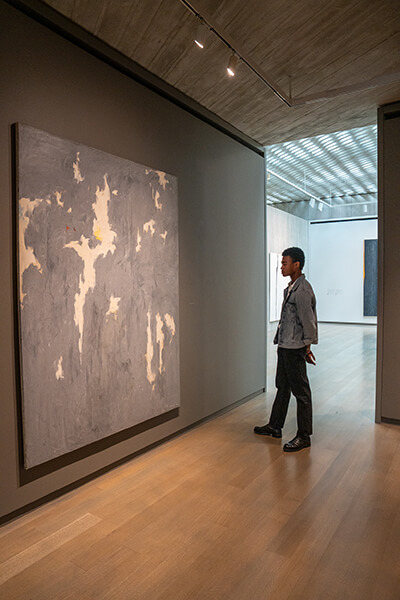 A young man wearing a jean jacket looks at a large gray and blue abstract painting