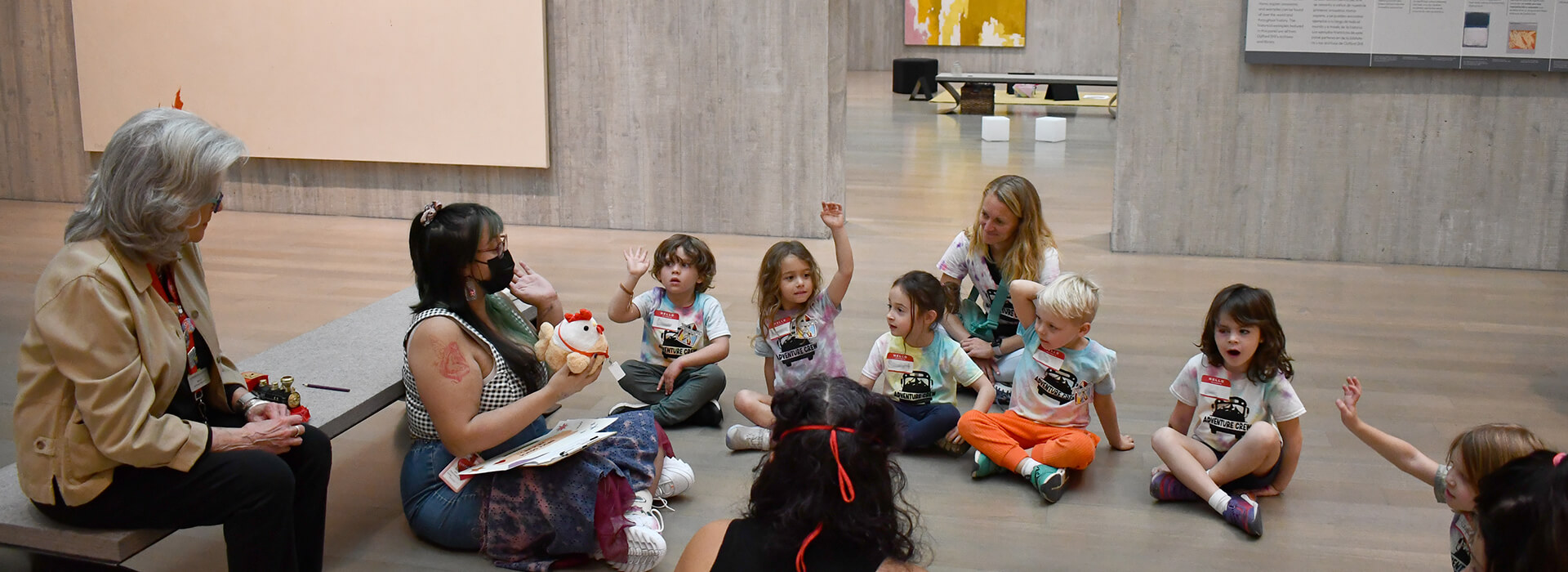 Pre-k students raise their hands while sitting on the floor of an art museum