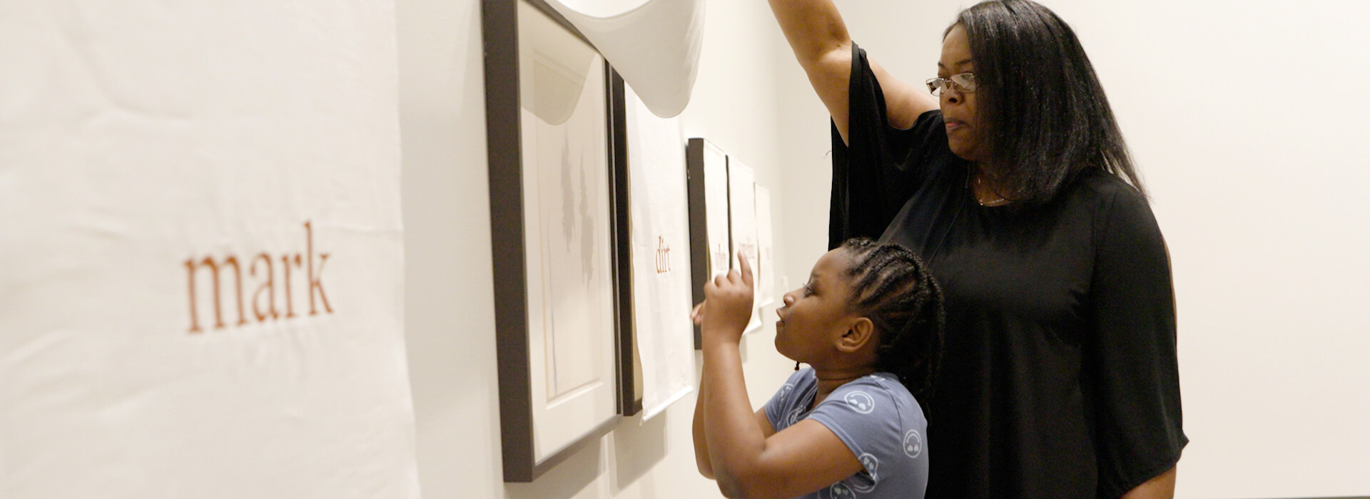 A woman lifts a curtain covering a pastel on paper artwork for her daughter to see