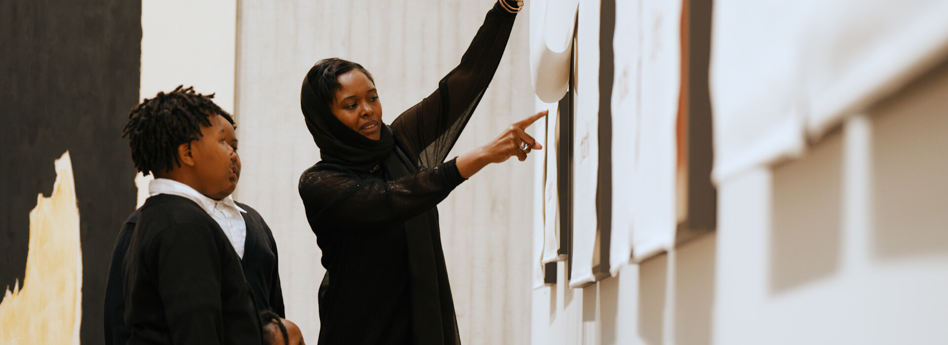 A woman lifts up a curtain and points at an artwork underneath to show her children