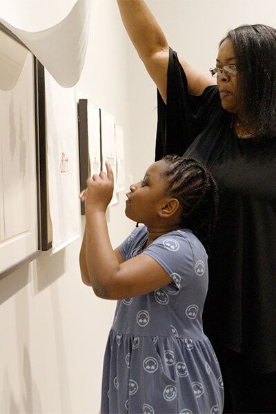 A woman lifts a curtain covering a pastel on paper artwork for her daughter to see