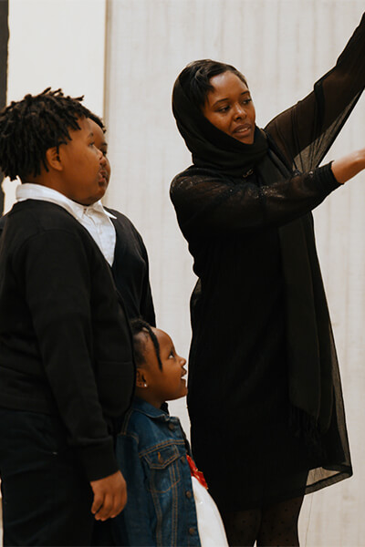 A woman lifts up a curtain and points at an artwork underneath to show her children