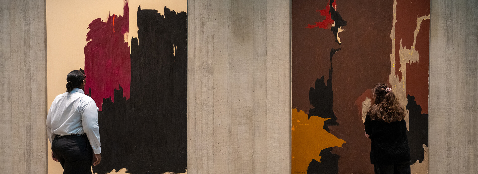 Two women look at abstract paintings hanging on a concrete wall