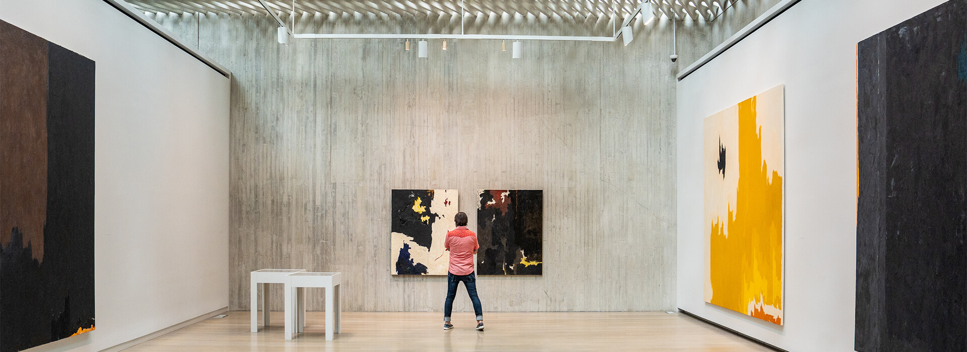 A man in a red shirt and jeans looks at abstract artworks on a wall