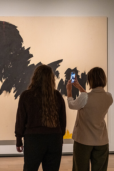 Two women look at a large abstract painting while one takes a photo with her phone