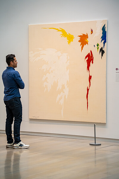 A young man looks at an abstract painting with large areas of bare canvas and fluttery colorful shapes in the upper right corner