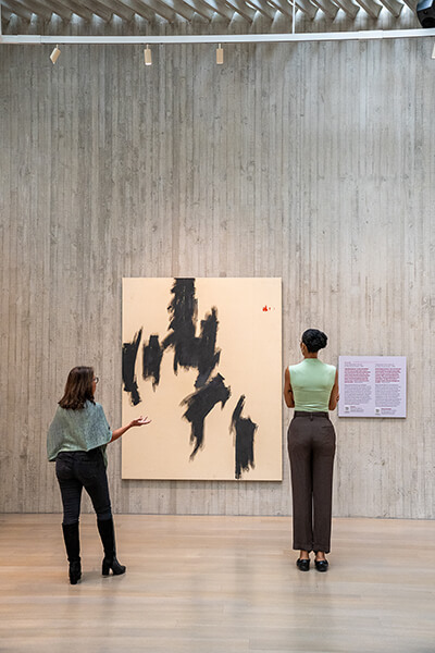 Two women look at an abstract painting with large areas of bare canvas and sections of jagged black paint