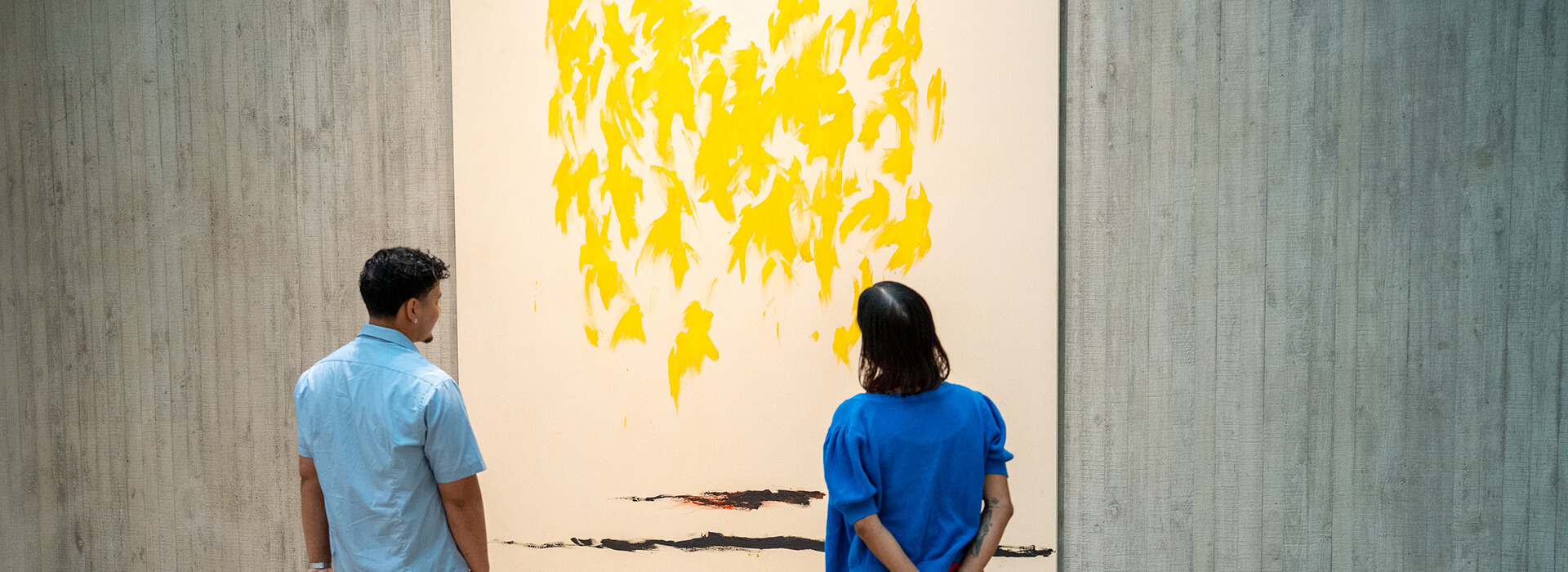 Two people look at a tall abstract painting with bare canvas, fluttery yellow forms, and horizontal dark lines