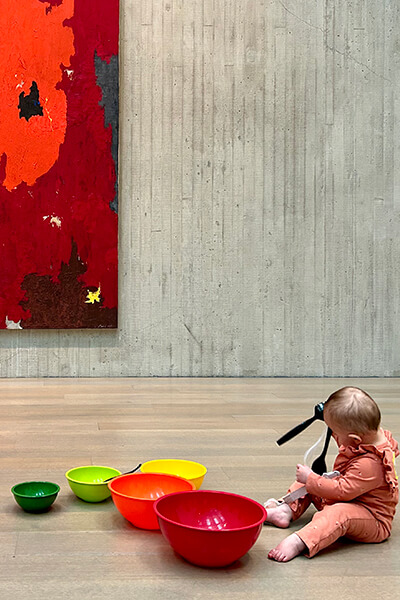 A baby bangs colorful plastic bowls on the floor of an art gallery