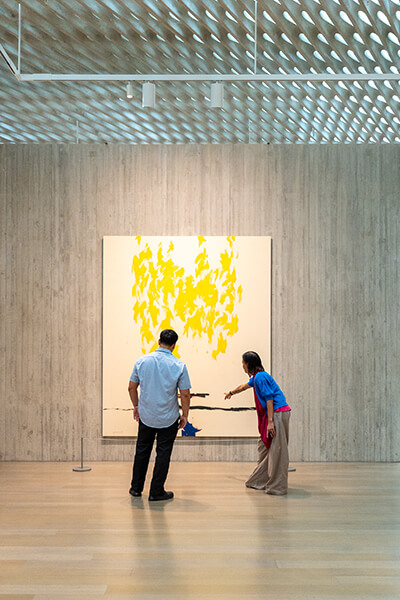 Two people look at a tall abstract painting with bare canvas, fluttery yellow forms, and horizontal dark lines
