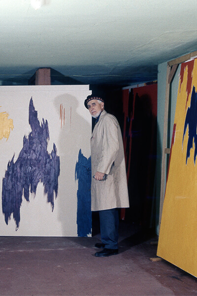 The images in this file come from 35mm slide photography of Clyfford Still posing in his basement work space at 312 Church Street in New Windsor, Maryland. The images depict Clyfford in a painting smock and beret, holding paintbrushes or posing nearby several of his later large-scale works.