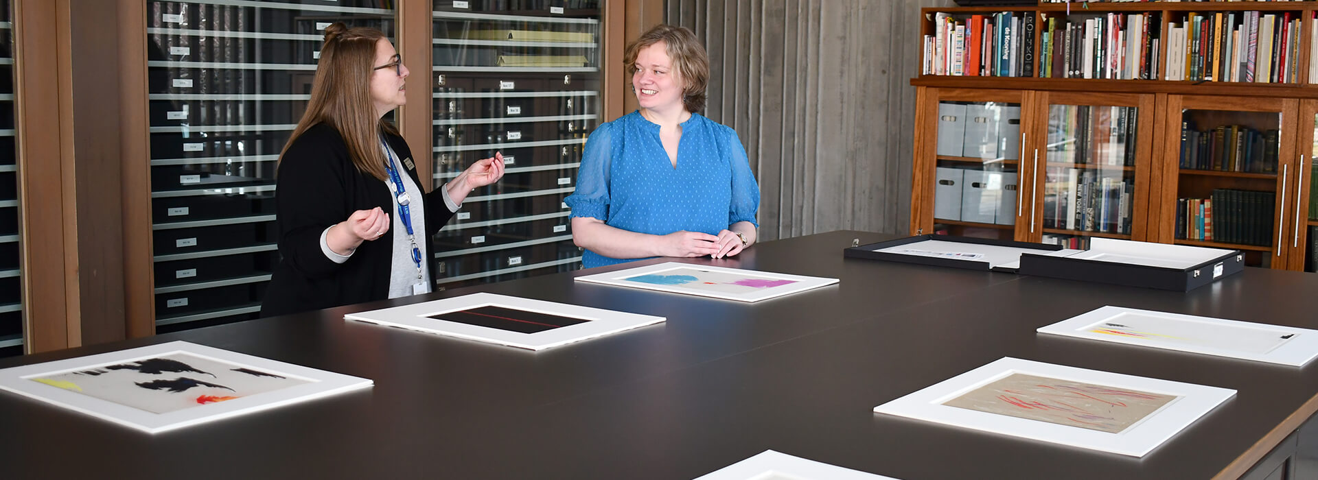 Two women talk inside the research lab at the Clyfford Still Museum with works on paper on the table in front of them