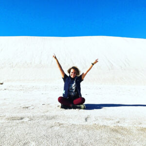 A person sits down on a sand dune and puts their hands in the air