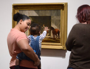 A woman holds a baby that reaches towards art