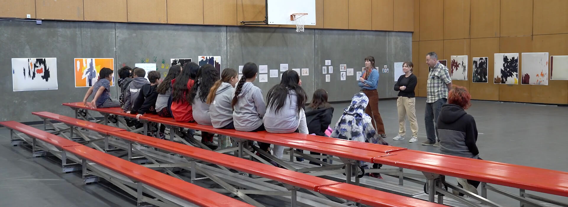 Three adults stand in front of a group of students on bleachers in a gym