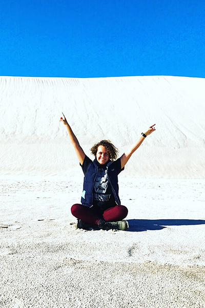 A person sits down on a sand dune and puts their hands in the air