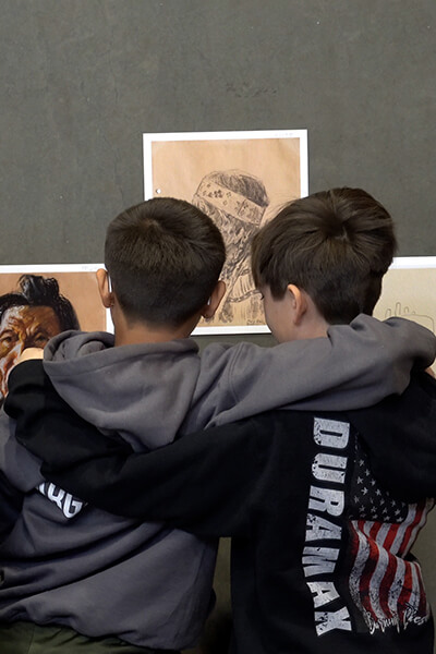 Two boys have their arms around each other's shoulders as they look at art