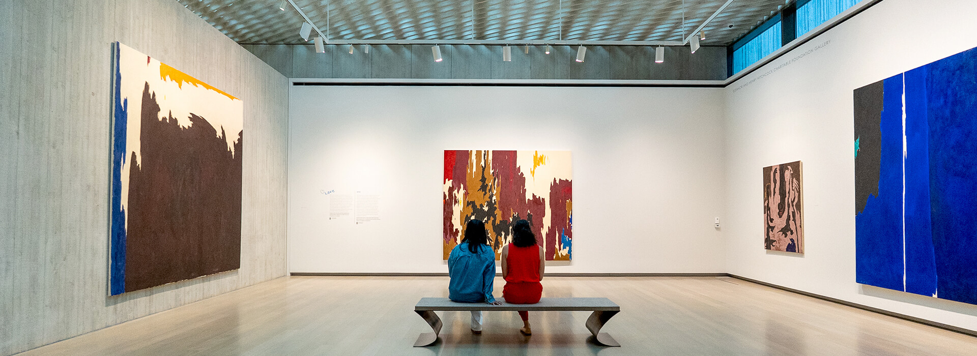 Two women sit on a bench and look at a large abstract painting
